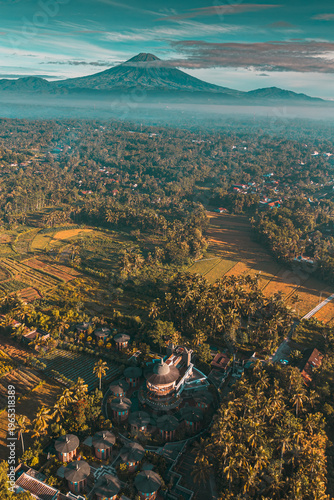 An aerial view reveals a grand mountain peak shrouded in clouds, overlooking a vibrant tropical landscape dotted with palm trees, terraced fields, and a unique circular resort under a serene sky.