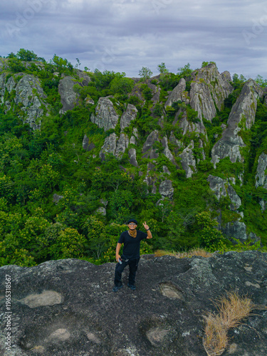 Aerial photo of a male adventurer on a rocky mountaintop. Drone photo of a solo traveler on a pristine & beautiful ancient rocky hilltop with a backdrop of other rocky mountains covered in green trees