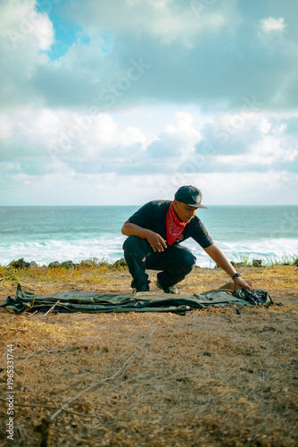 A male adventurer prepares to set up a tent alone by the sea. Solo camping activity on a hilltop near the beach with a view of the vast ocean and clouds. Male adventurer, tent, and ocean all at once.