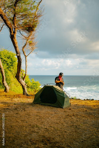 A male adventurer prepares to set up a tent alone by the sea. Solo camping activity on a hilltop near the beach with a view of the vast ocean and clouds. Male adventurer, tent, and ocean all at once.