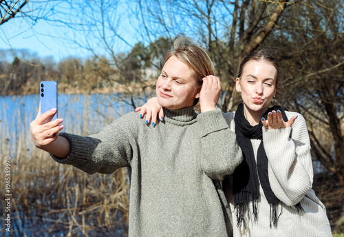 Two women have fun taking photos on a smartphone on a spring day in a park by the river.