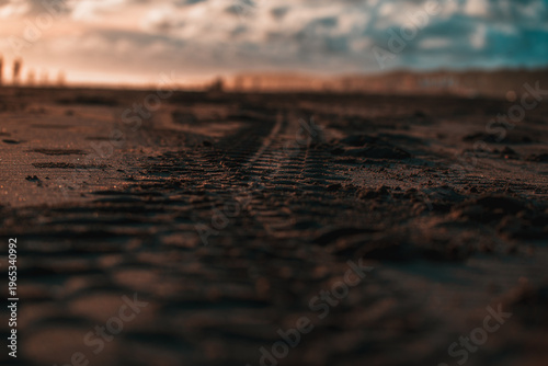 A close-up, shallow depth of field shot reveals intricate tire tracks in warm, sparkling sand with a blurred sunset or sunrise sky in the background.