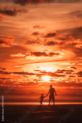 A heartwarming silhouette of a father and child holding hands while walking along a vibrant orange beach at sunset, with dramatic clouds reflecting the fiery sky.