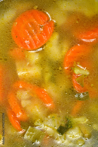 A vertical macro shot of homemade chicken soup with vegetables showing golden oil droplets on the surface and sliced carrots with broccoli in the broth