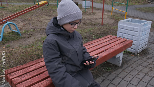 Cuadro en lienzo Child in winter clothing and glasses sitting on a park bench while intently scro