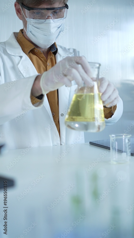 Fototapeta premium Male scientist wearing white protective gloves, mask and glasses is holding a yellow chemical solution inside an Erlenmeyer flask in a laboratory, vertical portrait. Science and medicine