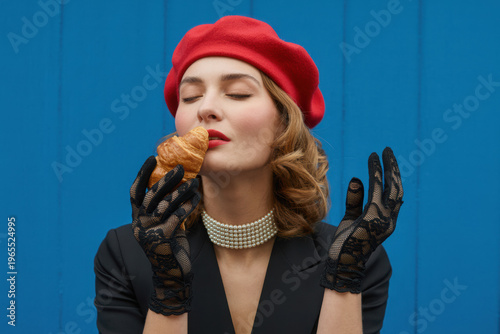 Fashion Portrait of Elegant Woman in Red Beret Eating Croissant on Blue Background