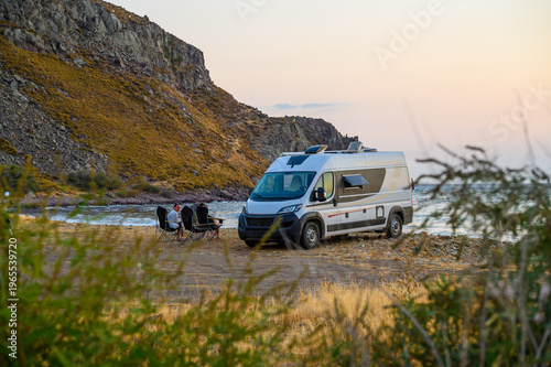 Family relax in folding chairs next to their modern campervan on a rugged beach in Limnos, Greece, enjoying a sunset view of the Aegean Sea during an off-grid motorhome road trip.
