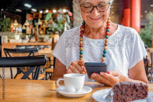 Smiling senior woman relaxing for a break at coffee shop with chocolate cake and coffee cup. Carefree elderly woman with eyeglasses and necklace using mobile phone