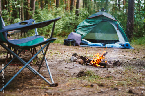 A cozy campsite in the woods featuring a small campfire, folding chairs, and a green tent under forest trees.