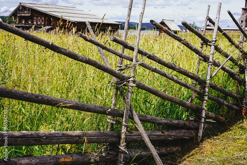 Fotografija A rustic wooden log fence borders a tall grass field with traditional timber houses under a cloudy sky