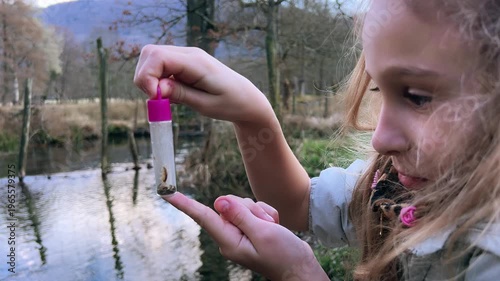 Little girl closely observing a small insect in a transparent collection tube near a pond in a natural park setting. Curious childhood nature exploration scene outdoors