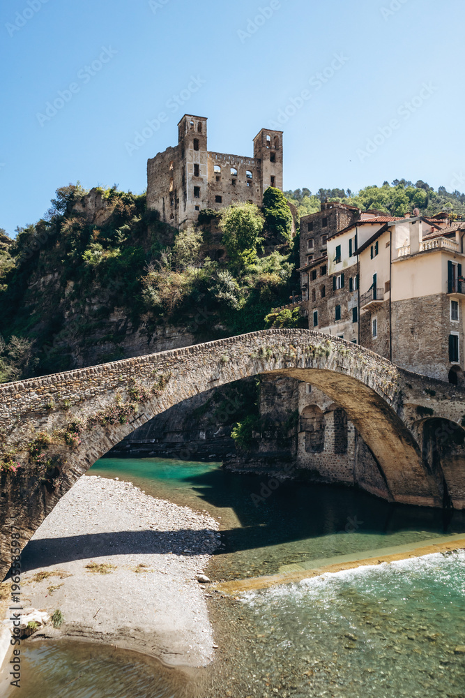 Obraz premium Historic stone bridge Ponte Vecchio over the clear waters of the Nervia river with the ruins of Doria Castle on the hill and traditional houses, sunny day in Dolceacqua, Liguria, Italy