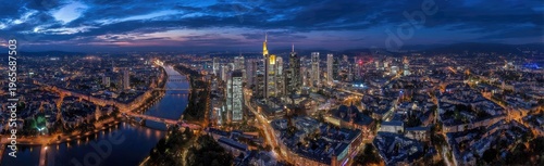 Aerial panorama of a city at dusk, with illuminated buildings, a river, and a colorful sky