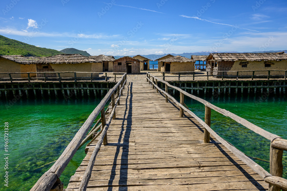 Fototapeta premium A long wooden boardwalk leads directly into the reconstructed Bay of Bones prehistoric stilt village, with transparent turquoise Lake Ohrid water glistening under a blue sky in North Macedonia.