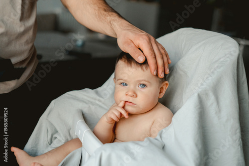 Soft light glows on a tender moment. Father's hand rests gently on a baby's head. The infant gazes calmly, fingers near lips. The baby's eyes hold quiet curiosity and trust