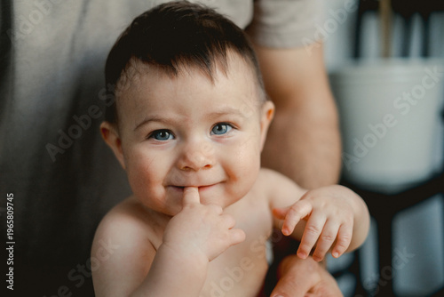 Cute baby smiles with finger to lips. Blue eyes sparkle with innocent joy. Soft lighting highlights baby's round face. Parent's arm supports baby gently. Perfect for parenting, family
