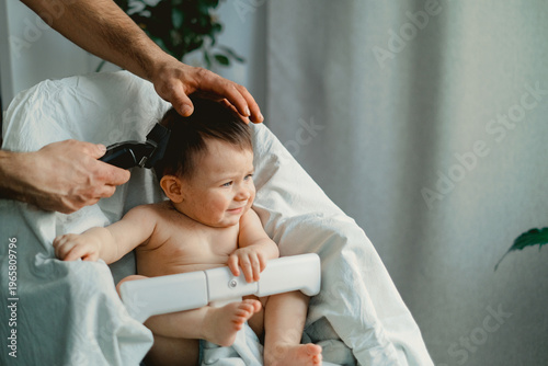 Tiny hands hold a baby's head gently. Curious eyes gaze forward, slightly puzzled. Adult hands guide a clipper with care. White towel wraps around baby's body softly. Soft lighting enhances calm