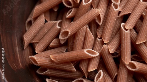 Raw wholemeal penne rigate pasta with rough ridged texture. Brown macaroni tubes scattered in a dark wooden bowl. Rotation. Top view
