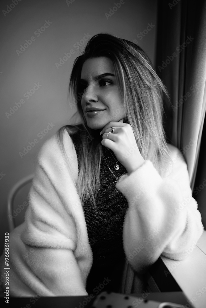 Naklejka premium Female model in a white coat and black thigh-high boots poses on a bar stool inside a modern cafe with a stylish interior and soft lighting. Black and white photo.