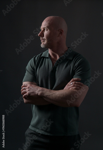 Confident mature bald man in dark green polo shirt looking aside on dark background, studio portrait with dramatic lighting