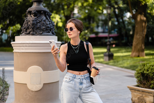stylish woman with short haircut walking in street with coffee wearing black top, jeans and sunglasses accessories