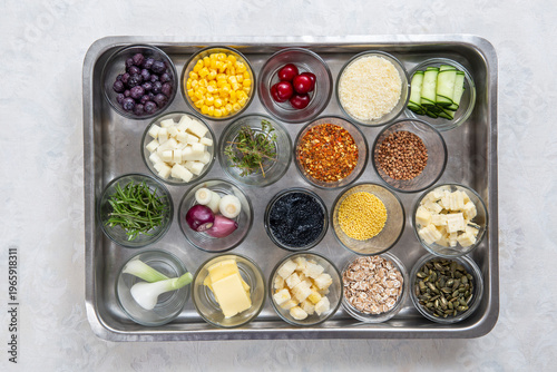 A metal tray with many small glass containers filled with a variety of prepared cooking ingredients such as vegetables, cheese, grains and herbs.