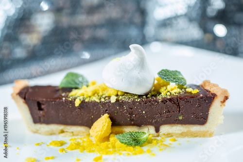 A piece of chocolate tart with a crispy base, decorated with white sprinkles, mint leaves and yellow crumbs on a white plate.