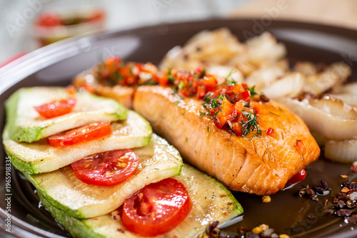 Grilled fish fillet served on a dark plate with a side dish of grilled zucchini with tomatoes and fried onions.