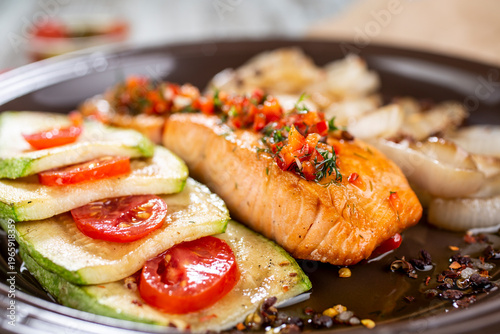Grilled fish fillet served on a dark plate with a side dish of grilled zucchini with tomatoes and fried onions.