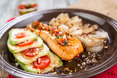 Grilled fish fillet served on a dark plate with a side dish of grilled zucchini with tomatoes and fried onions.