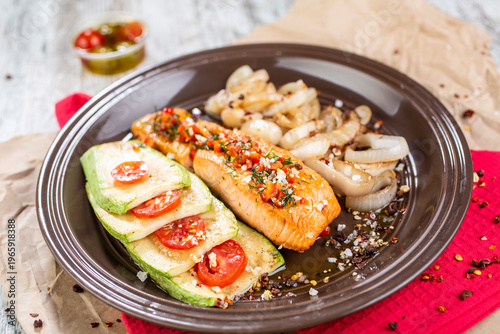 Grilled fish fillet served on a dark plate with a side dish of grilled zucchini with tomatoes and fried onions.