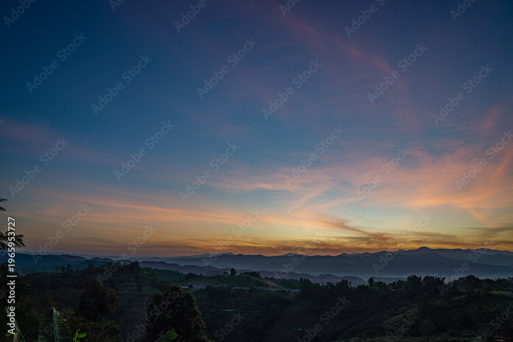 Fototapeta premium A breathtaking sunset over layered mountain ranges, with warm golden light fading into a deep blue sky, creating a serene and expansive natural landscape ideal for travel and nature themes.