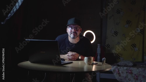 A man in black clothes sits at a table. He wears a baseball cap and glasses. He works remotely. Freelancer. Night. The lights are on. There's a laptop on the table. A ceramic mug. Home interior. Stora