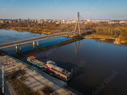 Aerial view of the Slasko-Dabrowski Bridge casting a stark shadow over the Vistula River, with the city skyline shimmering in the distance, Warsaw, Poland.
