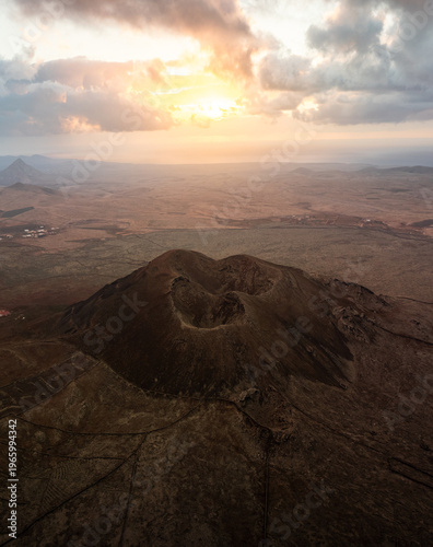 Aerial view of the volcano's dark slopes meeting the arid plains under a luminous sky, creating a mesmerizing contrast of light and shadow, Fuerteventura, Canarias, Spain.