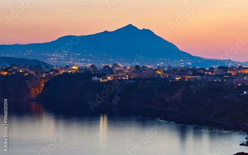 Long exposure evening photo of Procida with illuminated buildings on cliff above calm Mediterranean sea. Silhouette of Ischia island rises in background under sunset gradient sky