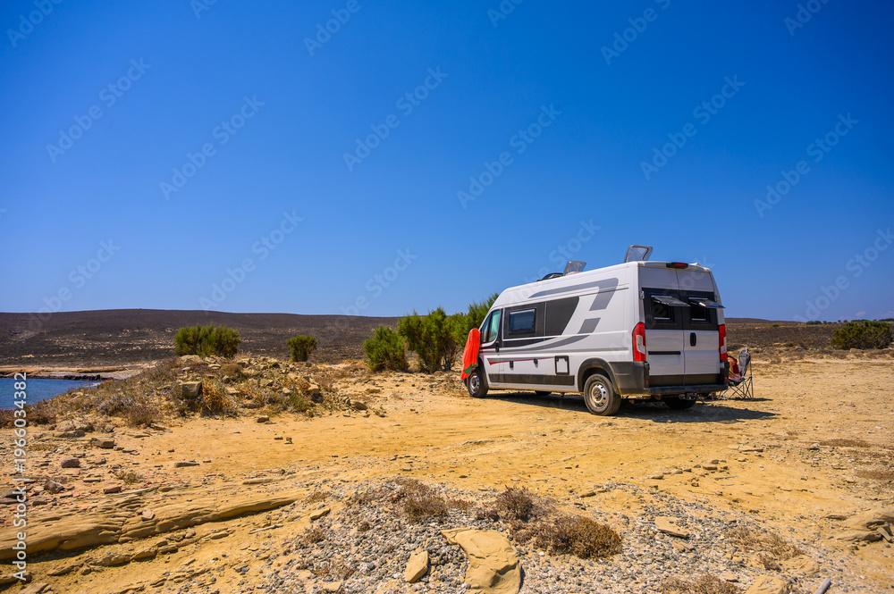 Obraz premium A family of travelers relaxes in folding chairs next to their modern campervan on a rugged beach in Limnos, Greece, enjoying a sunset view of the Aegean Sea during an off-grid motorhome road trip.