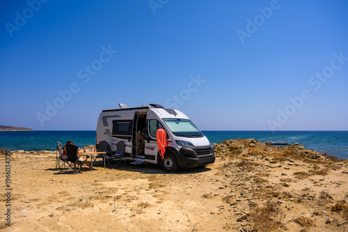 A family of travelers relaxes in folding chairs next to their modern campervan on a rugged beach in Limnos, Greece, enjoying a sunset view of the Aegean Sea during an off-grid motorhome road trip.