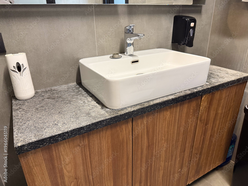 Fototapeta premium Modern ceramic sink with chrome faucet installed on stone countertop in public restroom interior. Hygiene maintenance, handwashing routine, sanitation infrastructure and facility management concept