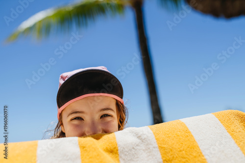 Smiling child peeking over beach towel under palm tree.
