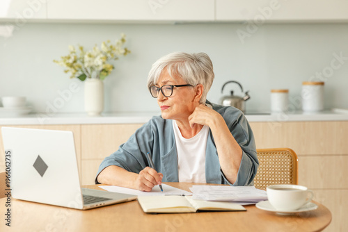 Middle aged senior woman using laptop computer writing notes at home. Focused mature old businesswoman work in home office making written records doing online work. Mature boss professional worker