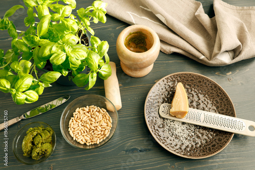 Traditional Genovese pesto preparation with fresh ingredients on wooden background. High angle shot of marble mortar with pestle, fresh basil, pine nuts, olive oil and parmesan cheese with grater for