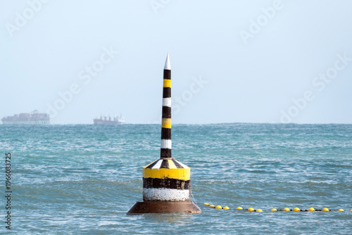 Historic black, white and yellow pylon located at Cottesloe Beach with container ships on the horizon, Perth, Western Australia.