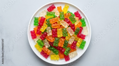 An overhead flat-lay of colorful gummy bear candies in red, orange, yellow, green, and clear varieties piled abundantly on a clean white ceramic plate against a smooth light grey studio surface, the