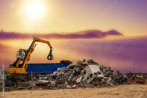 Grapple excavator moving metallic waste at an industrial recycling facility under a vibrant sky