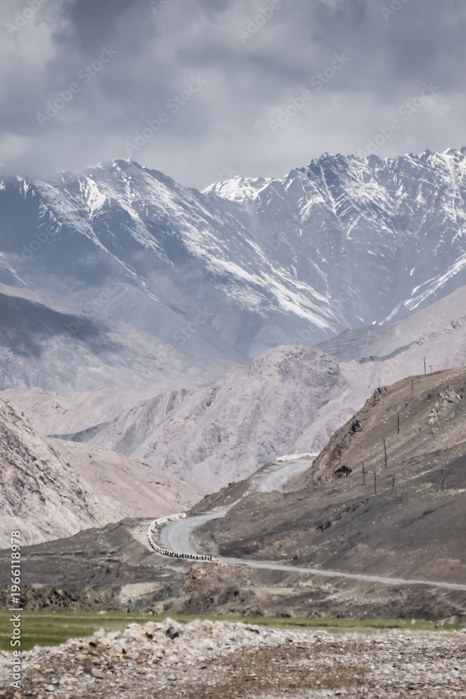 Fototapeta premium The Pamir Highway, a paved road in a valley of rocky mountains, with high mountain ranges and peaks covered with snow and glaciers in the background, a landscape in the Tien Shan Mountains