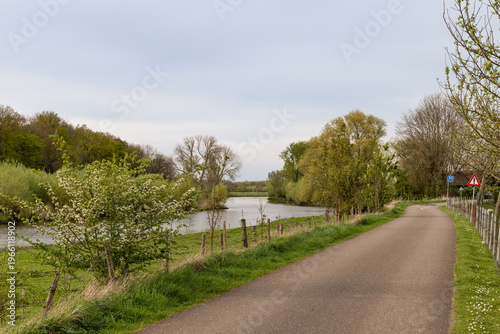 Scenic bicycle path along the Linge river in the Betuwe, Netherlands. Spring landscape with green trees, water, and rural road.