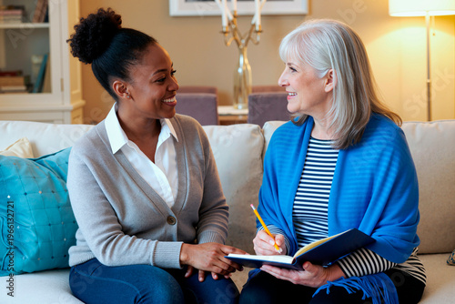 Middle aged Black woman smiling while talking with senior Caucasian woman holding notebook and pencil, both sitting on sofa engaging in friendly conversation in living room
