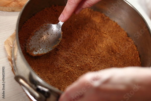Leveling ground cookies mixture with spoon in baking pan. Making frozen strawberry cheesecake series.